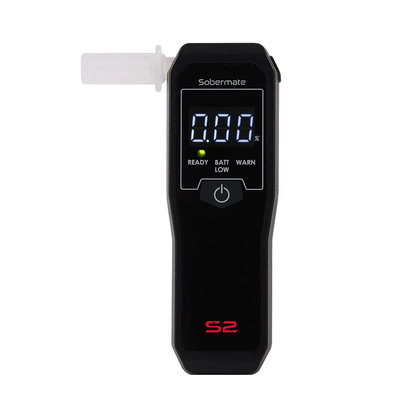 Tiny matte-black Sobermate S2 breathalyser next to a standard credit card for scale; device features a bright colour OLED screen showing “0.00” result, single button, and fold-out blow tube mouthpiece